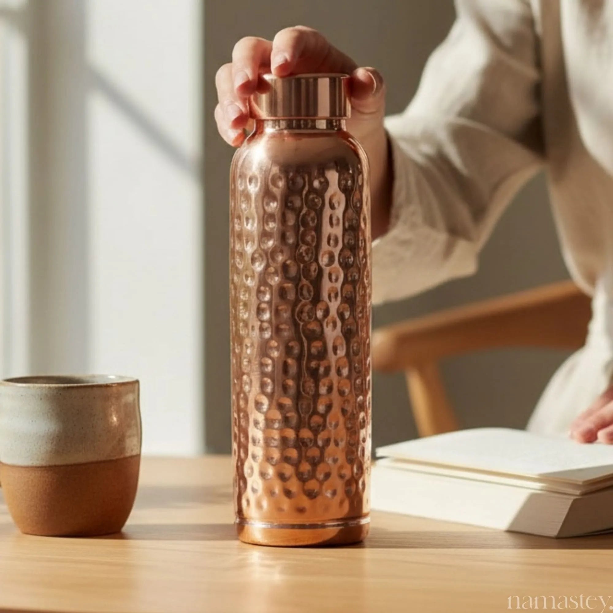Hand placing the classic hammered copper bottle on a desk beside a mug and notebook